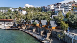 A coastal hotel with multiple wooden cabins, a stone pathway leading to the beach, and a backdrop of modern buildings and trees. - Roseira Bungalov (Milas)