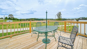 Una terraza de madera con una mesa y sillas de metal verde, un farol de metal, y vista a un área con césped y árboles.