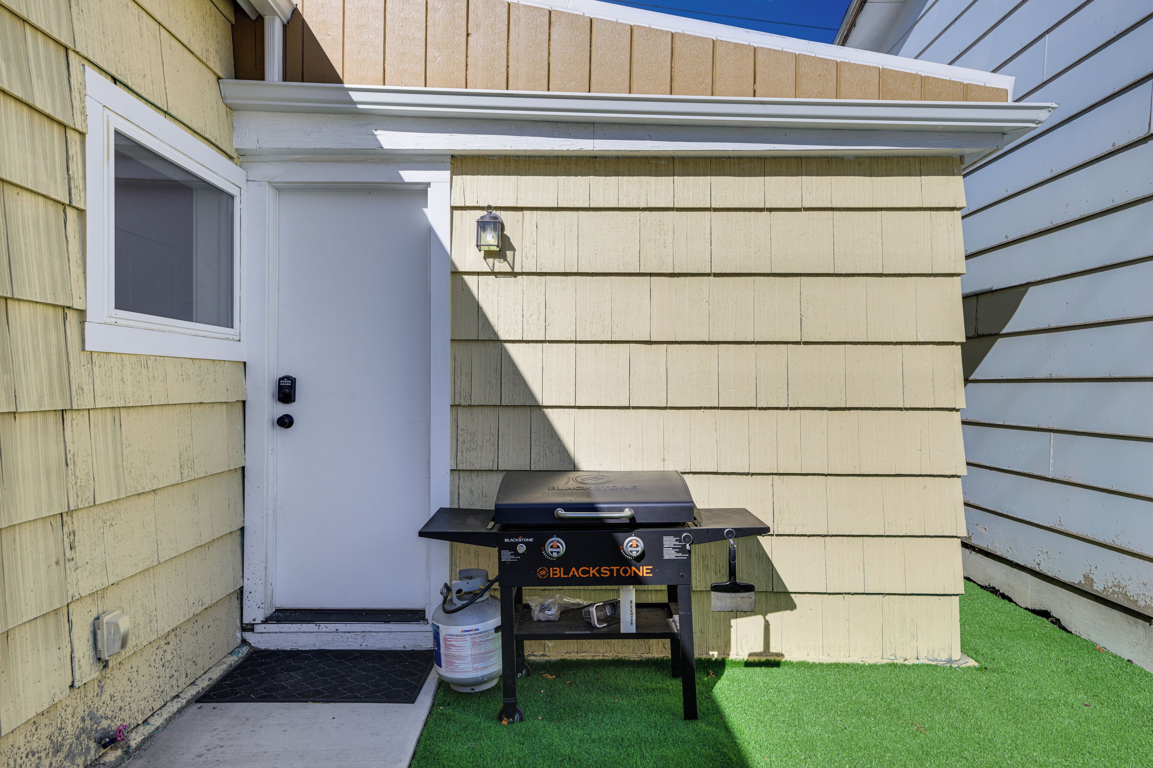 En svart utomhusgrill med Blackstone-logotypen, placerad på konstgräs bredvid ett hus med beige fasadpanel och en vit dörr.