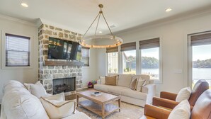 A living room with a stone fireplace, a large flat-screen TV, a beige sofa, a wooden coffee table, and a view of a lake. - Waterfront Home in Alex City, Near Auburn (Alexander City)