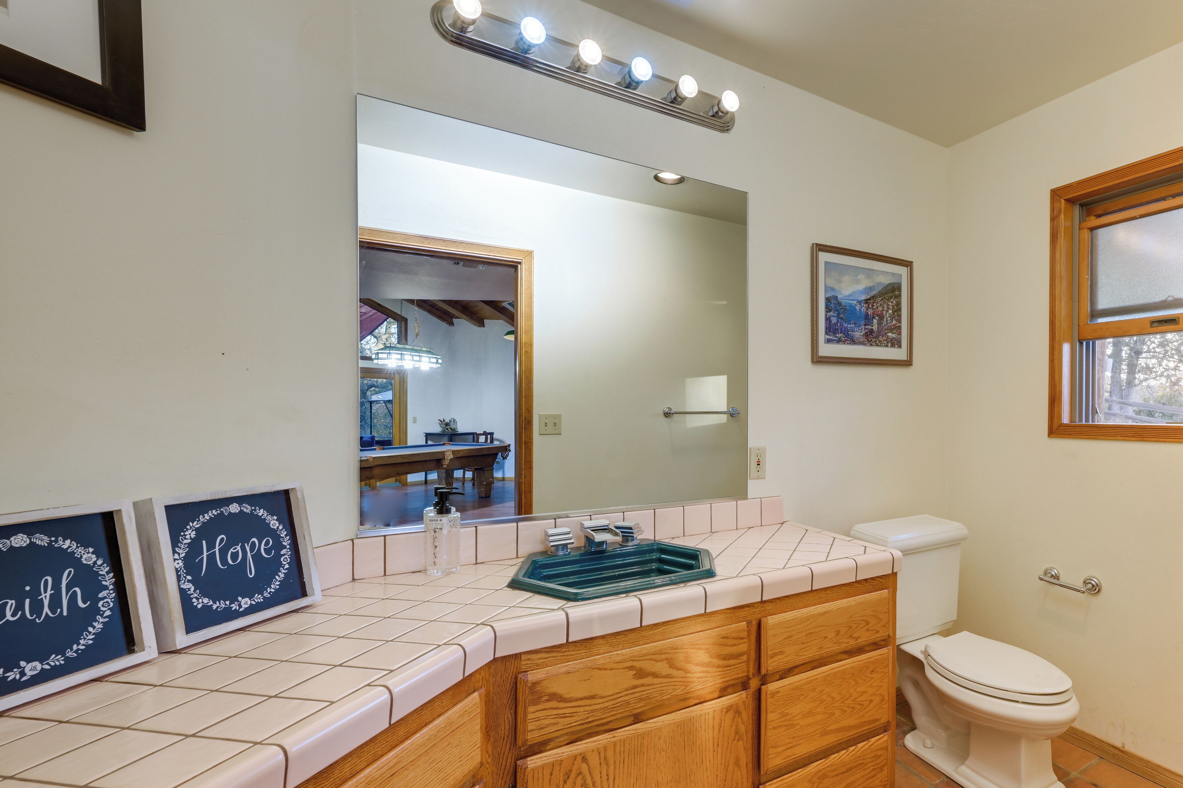 A bathroom with a tiled vanity, a mirror with lights, framed pictures, and a toilet.