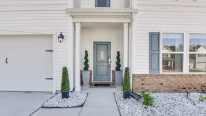 Une maison à deux étages, avec une façade blanche, une porte d’entrée verte et une porte de garage. Deux plantes en pot sont placées de chaque côté de l’entrée.