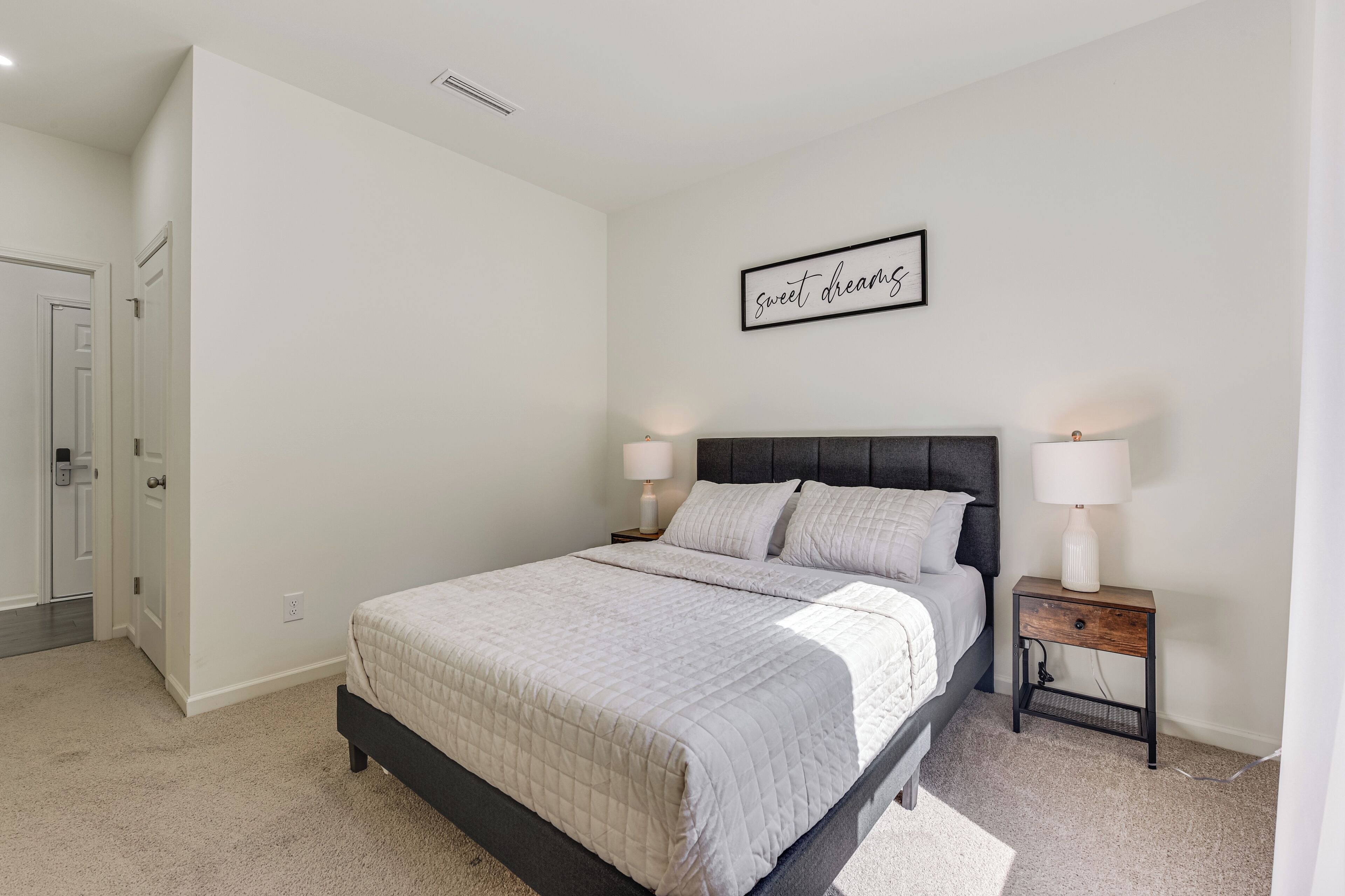 A bedroom with a bed, two bedside tables with lamps, a door, and a wall with a framed sign.