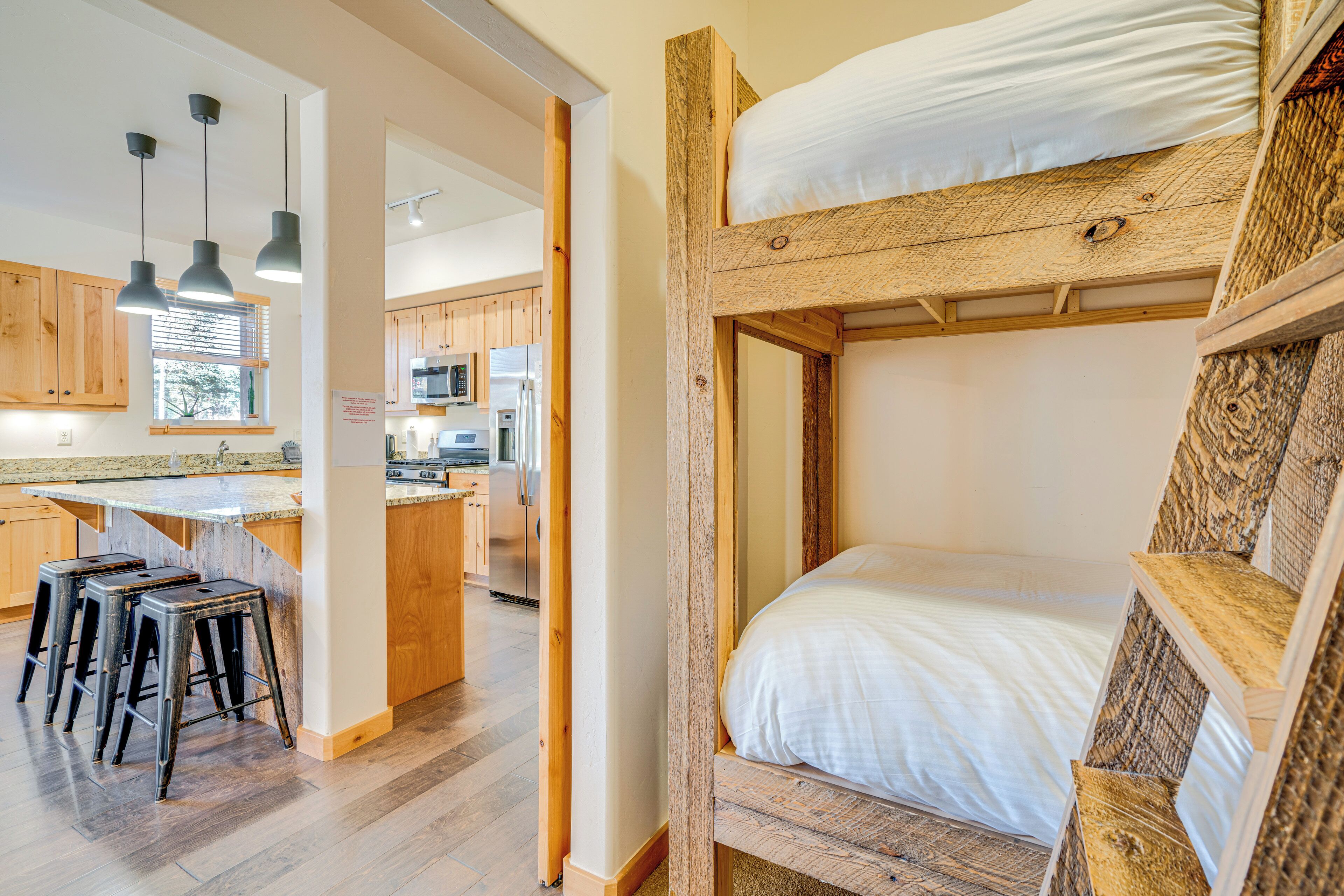 Una cama tipo litera con cabecera de madera, una zona de cocina con armarios de madera y un espacio con taburetes de bar.