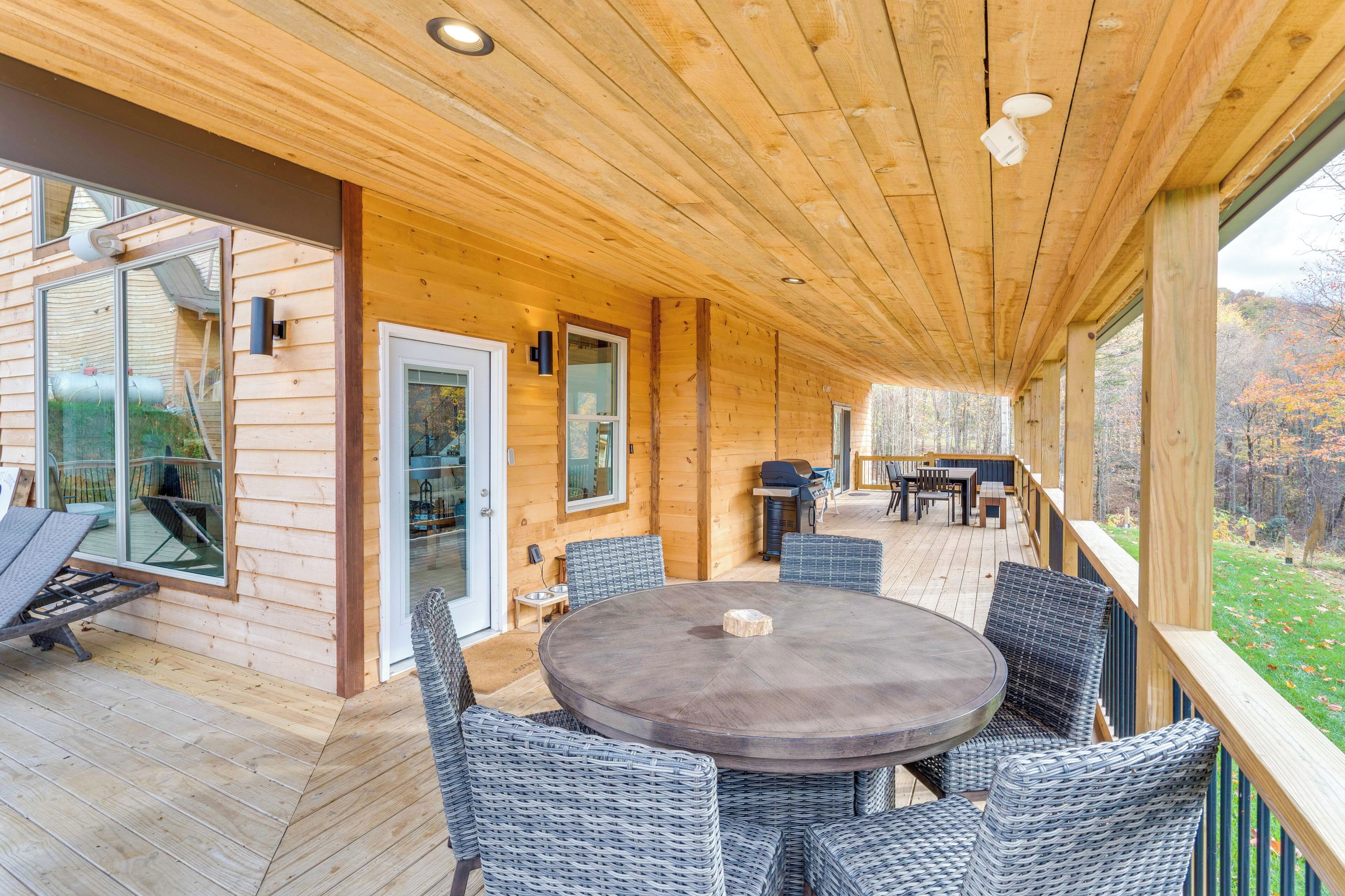 A wooden deck with a round table and wicker chairs, a barbecue area, and glass doors leading to a patio.