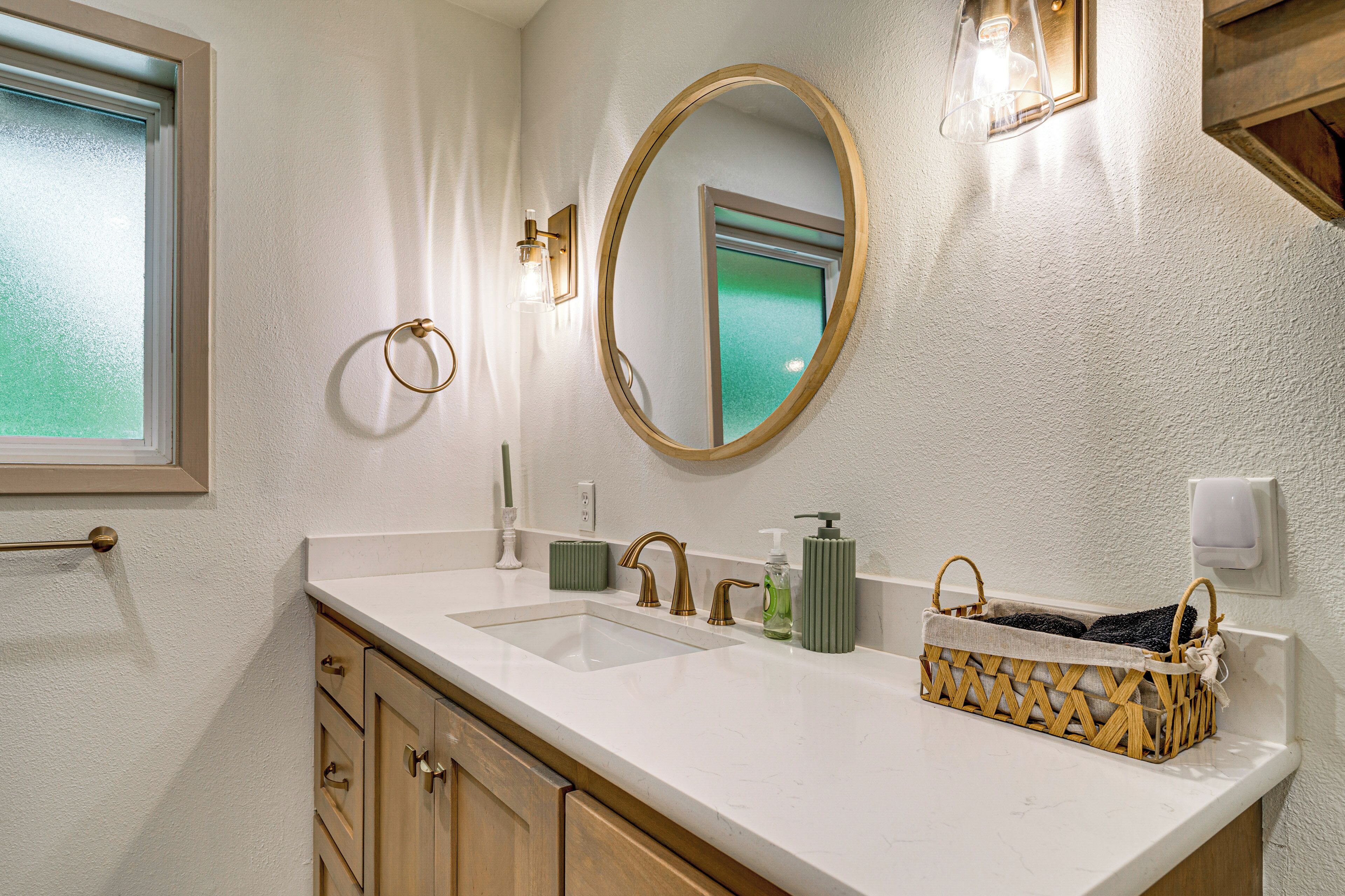 A bathroom with a white sink, gold faucets, a round mirror, and a wooden basket on the counter.