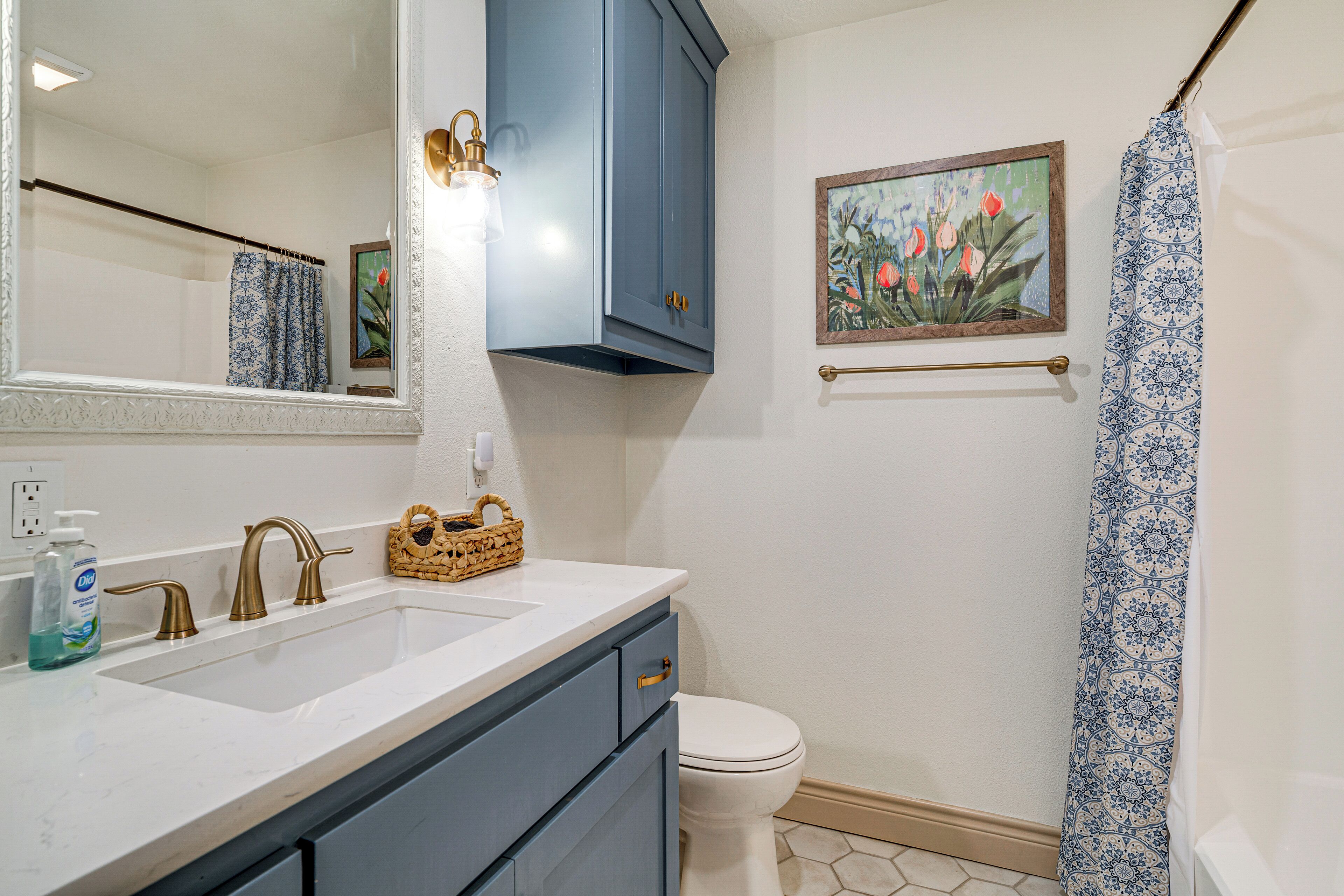 Een badkamer met een wit wastafel, twee gouden kraanwerk, een blauwe kast, een wit toilet, een douchegordijn en een gepoft bloemstillevaan de muur.