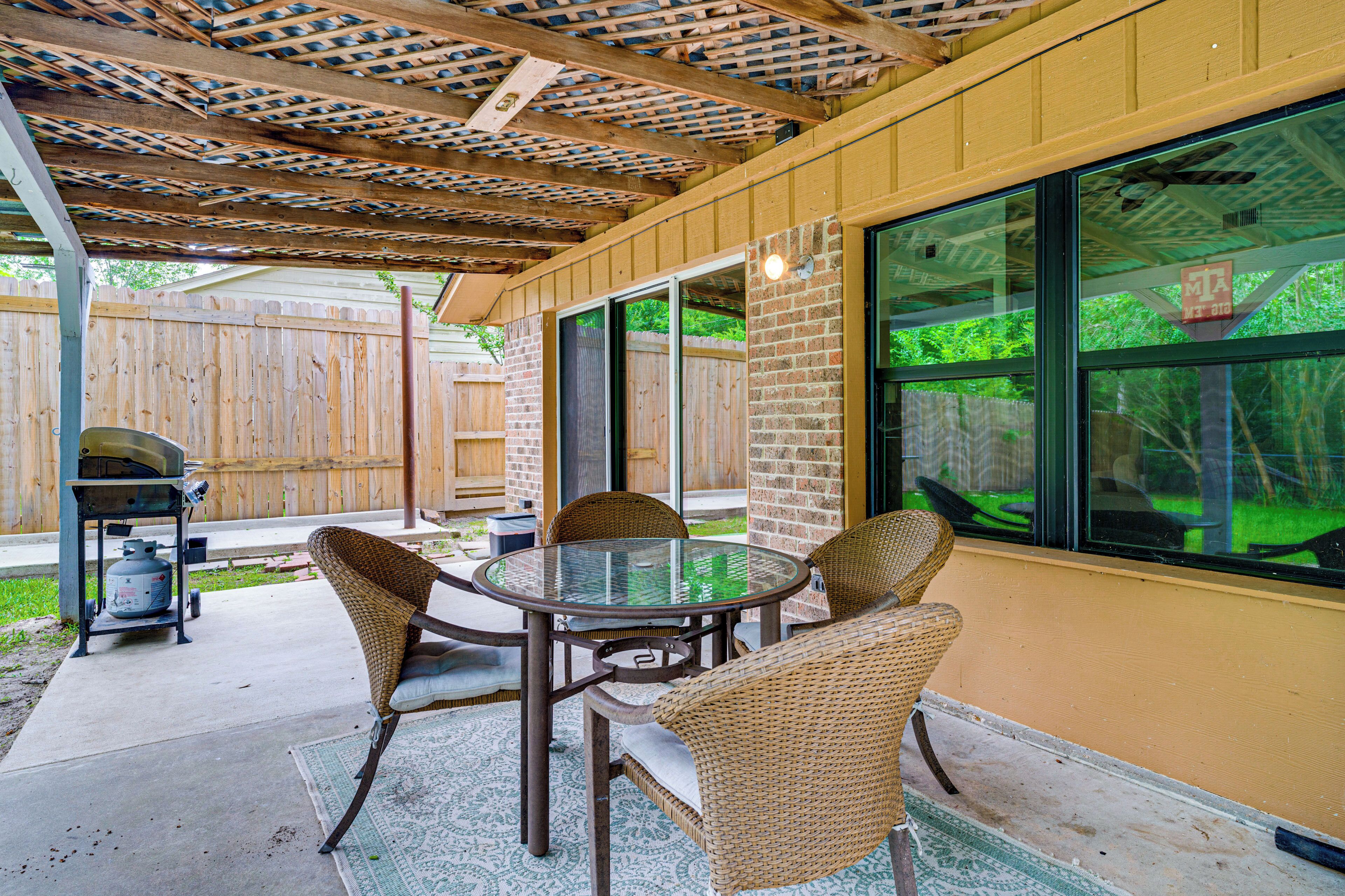 Een terras buiten met een ronde glazen tafel, rieten stoelen, een barbecue en uitzicht op een houten schutting en groen.