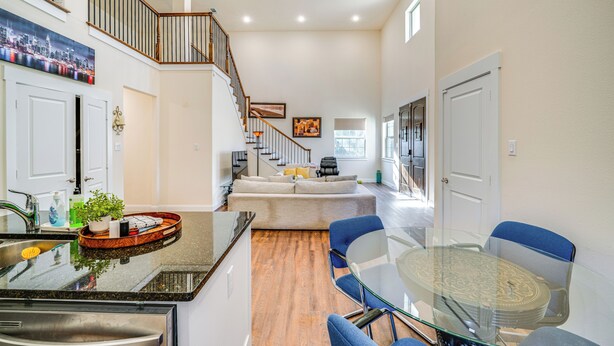 A modern kitchen with a glass dining table, blue chairs, and a staircase leading to a loft area. - Walk to Historic Dtwn: Mckinney Family Retreat (McKinney)
