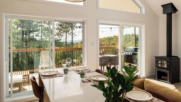 A dining area with a table set for four, large windows, a black wood stove, and a view of a deck and trees. - La Selva - Intimate getaway & river access (Sainte-Béatrix)