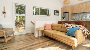A modern living room with a sofa, a small side table, and a view of a forest through large windows. - La Selva - Intimate getaway & river access (Sainte-Béatrix)