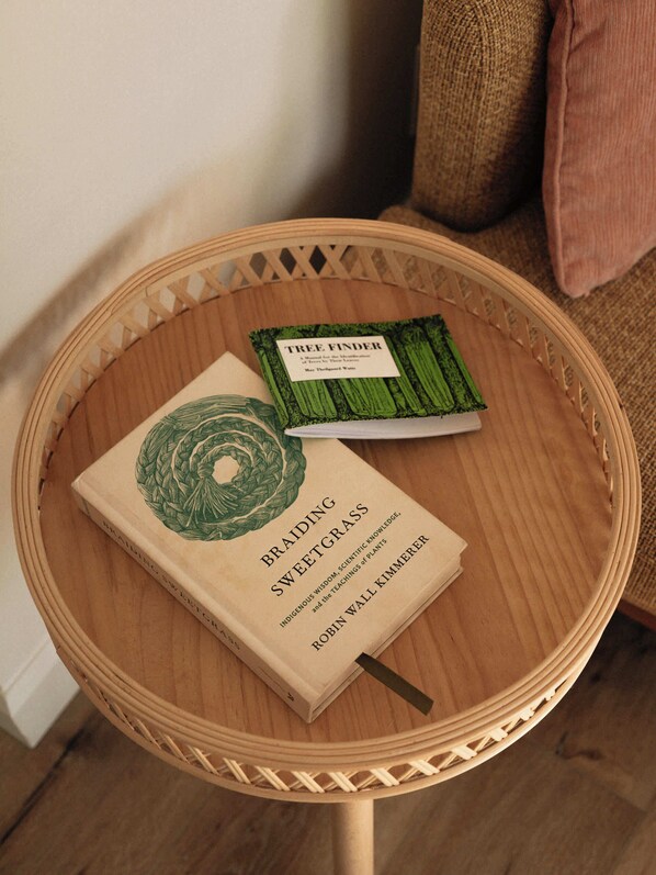 A round wooden table with a woven edge, holding two books titled 'Braidings Sweetgrass' and 'Tree Finder'.