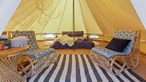 A cozy tent interior with a bed, two wicker chairs, a striped rug, and a table with a box and a bottle.