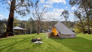 A tent set up on a grassy field with a fire pit nearby, surrounded by trees and a small building in the background.