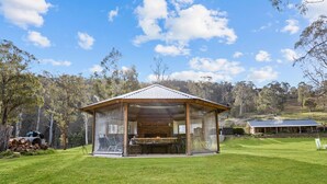 A covered outdoor barbecue area with a brick fireplace, surrounded by a grassy lawn and trees.