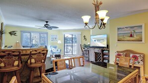 A spacious living room with a large glass dining table, wicker furniture, a ceiling fan, and a view of the outdoors through the sliding glass door. - Three bedroom Shipyard villa (Hilton Head)