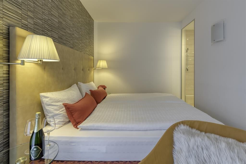A hotel room with a bed, two bedside lamps, a bottle of champagne, and a glass on the nightstand.
