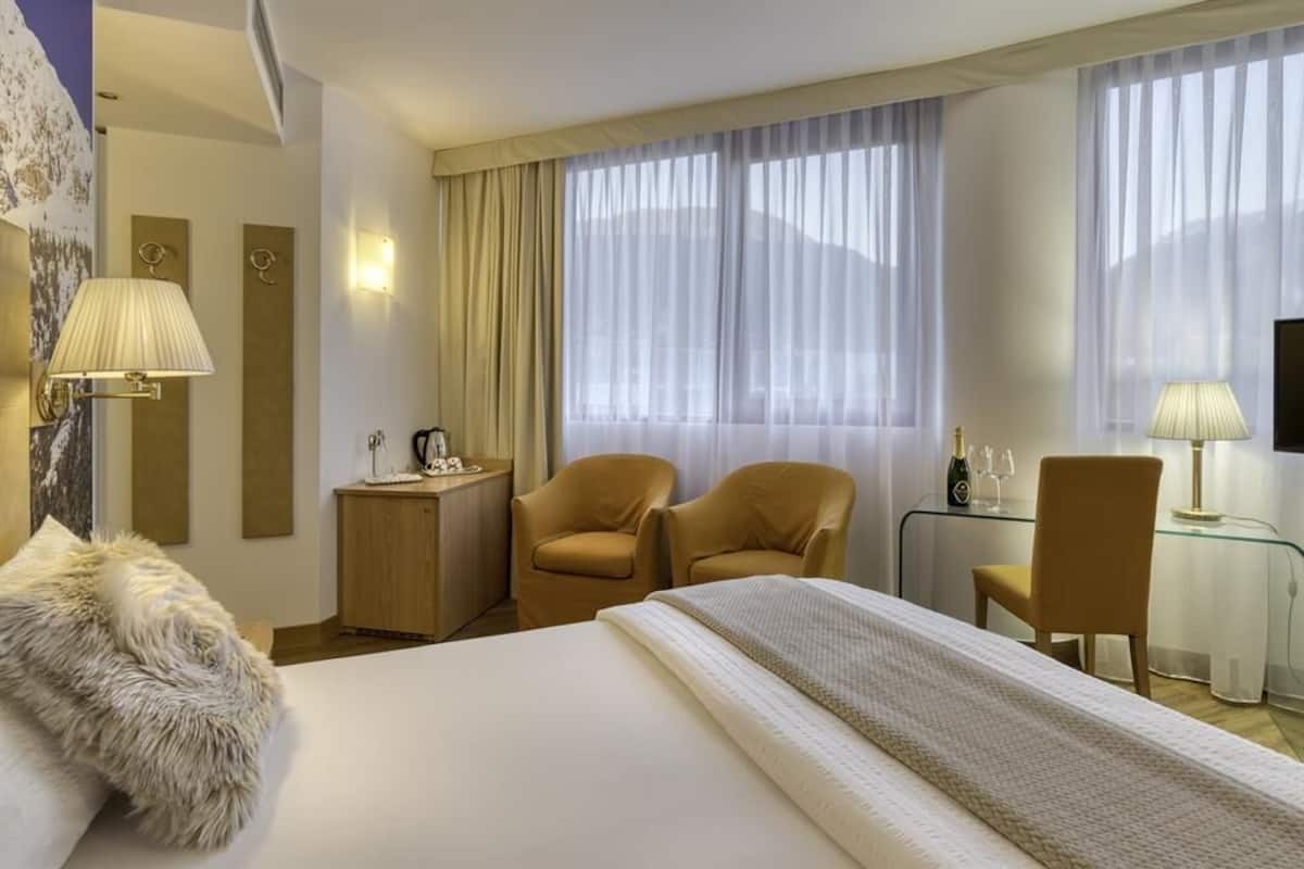 A hotel room with a large bed, two chairs, a desk with a lamp, a minibar, and a large window with curtains.