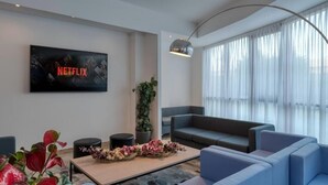 A modern living room with a large flat-screen TV displaying the Netflix logo, a sofa, a coffee table with decorative items, and a large window with sheer curtains. - Hotel Sporting Trento (Trento)