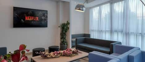 A modern living room with a large flat-screen TV displaying the Netflix logo, a sofa, a coffee table with decorative items, and a large window with sheer curtains.