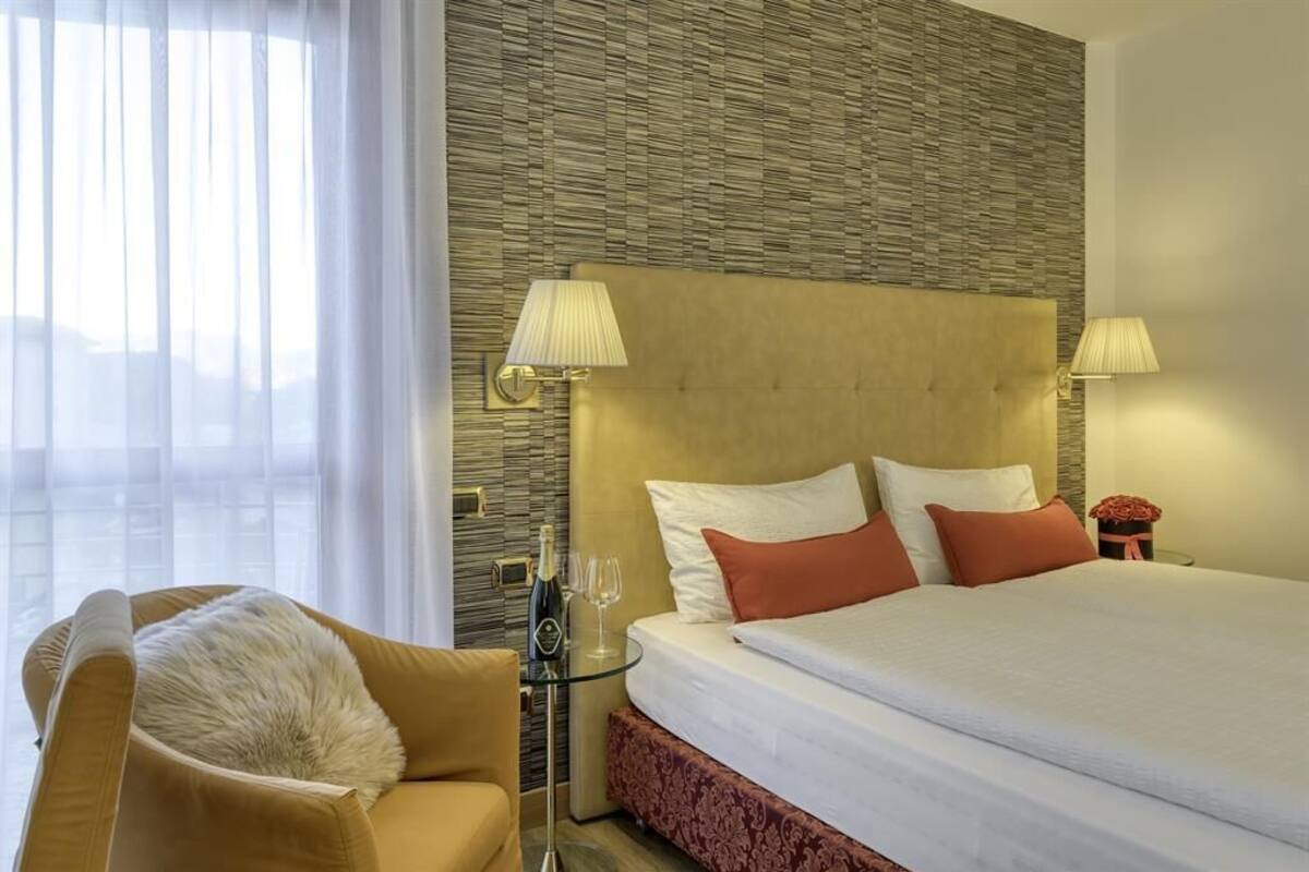 A hotel room with a large bed, two bedside lamps, a glass table with a bottle and glasses, a chair with a fur throw, and a view of the city through sheer curtains.