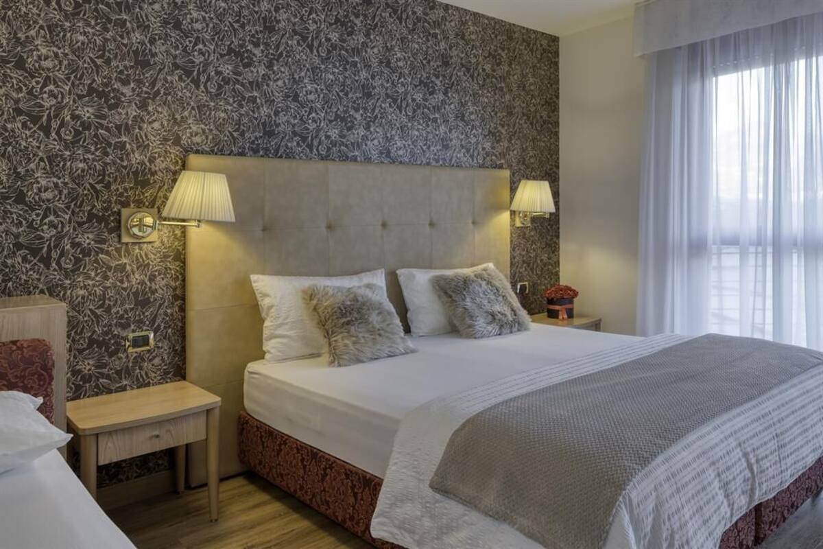 A hotel room with a large bed, two bedside lamps, a nightstand, and a floral-patterned wall.