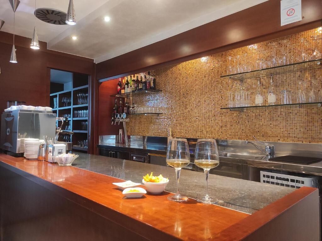 A bar area with a wooden counter, glasses, and bottles. There are shelves with glasses and bottles in the background.