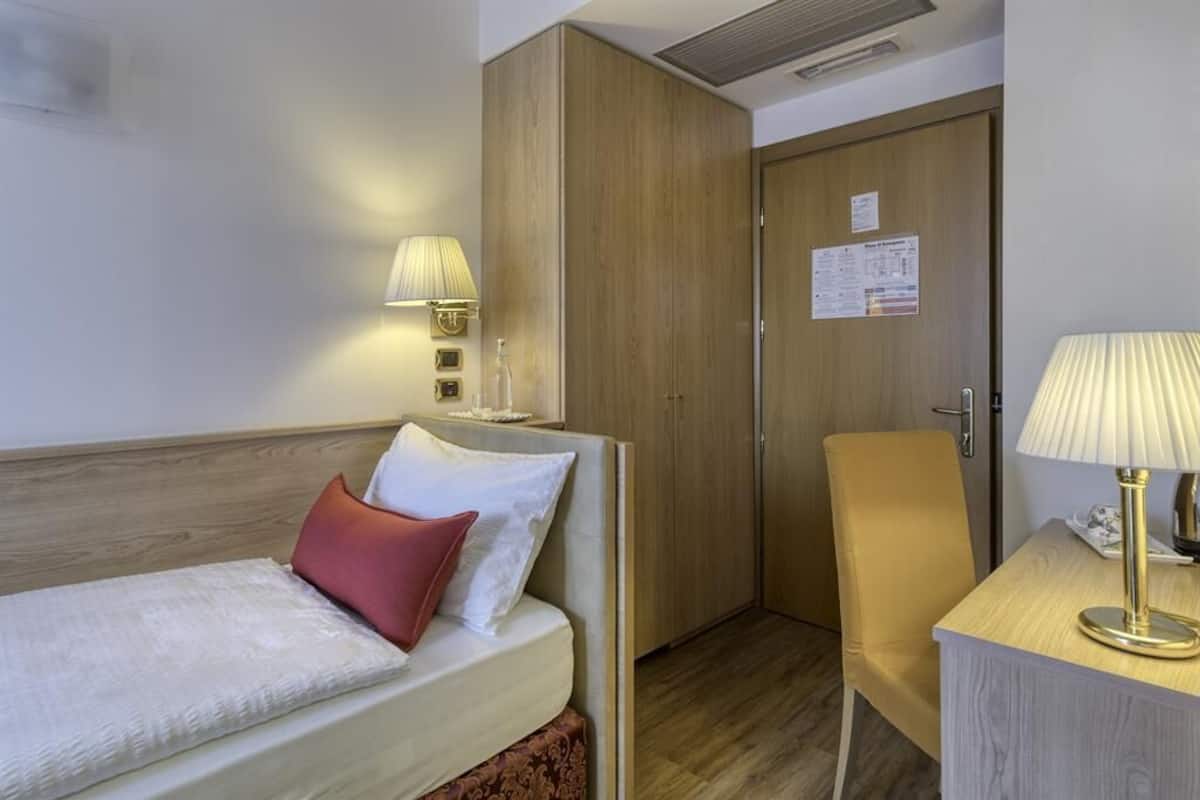 A hotel room with a bed, a wooden wardrobe, a desk with a lamp, and a door with a notice.