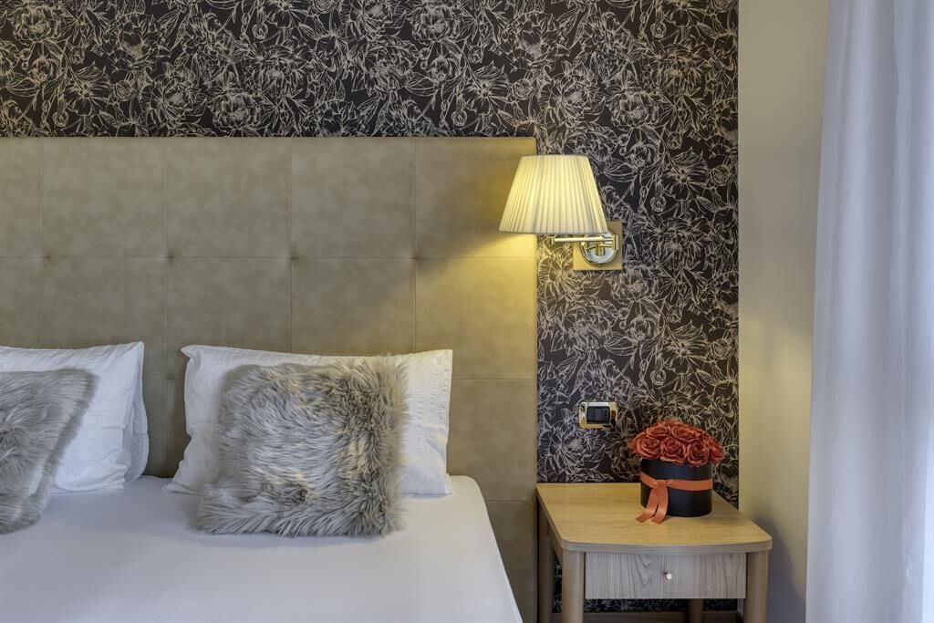 A hotel room with a tufted headboard, a bedside lamp, a small wooden nightstand with a flower arrangement, and a wall with a floral pattern.