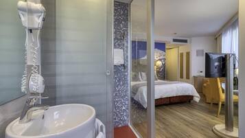 Hotelkamer met glazen douchecabine, wit wastafelmeubel, bed met witte dekbedovertrek, televisie en houten vloer.