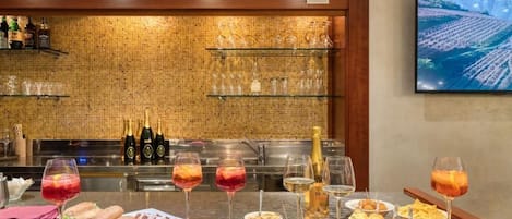 Zona de bar con salpicadero de azulejos, estantes con cristalería, botellas y una pantalla de televisión mostrando un viñedo.