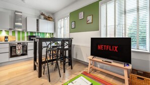 Et moderne køkken med spiseplads, et fjernsyn der viser 'NETFLIX', og et vindue med persienner.