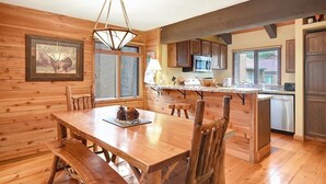 A wooden cabin interior with a dining table, chairs, a kitchen with a microwave, and a hanging light fixture. - Wildflower 62: Nice Condo, Close to the Green Line (Mammoth Lakes)