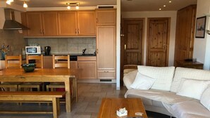 A modern living room with a wooden coffee table, a beige sofa, and a kitchen area with wooden cabinets and a microwave. - 3 Bedroom Apartment Nendaz, Switzerland. 5 mins walk to tracouet gondala. (Nendaz)