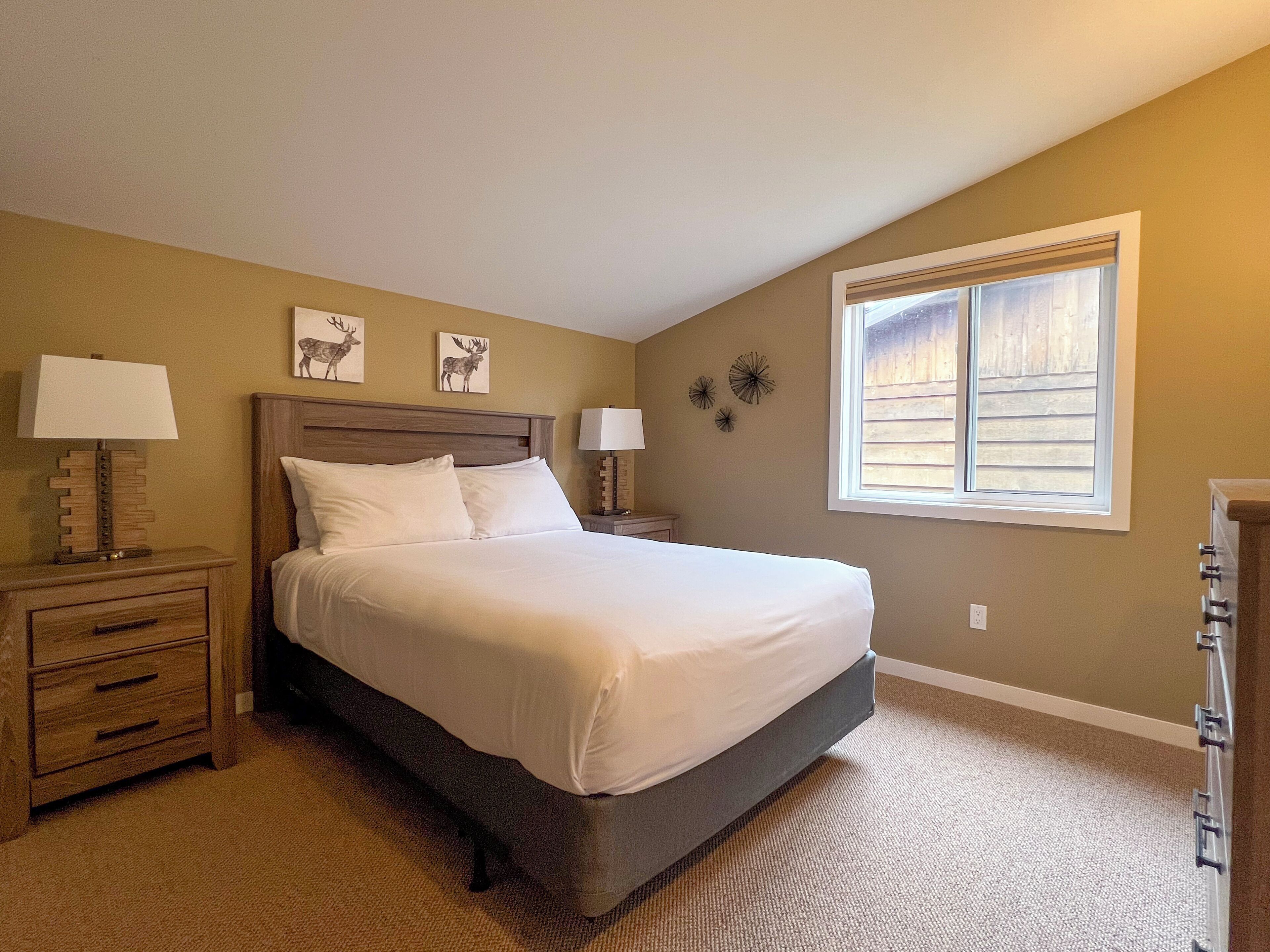 A bedroom with a large bed, two bedside tables with lamps, a dresser, and a window with blinds.