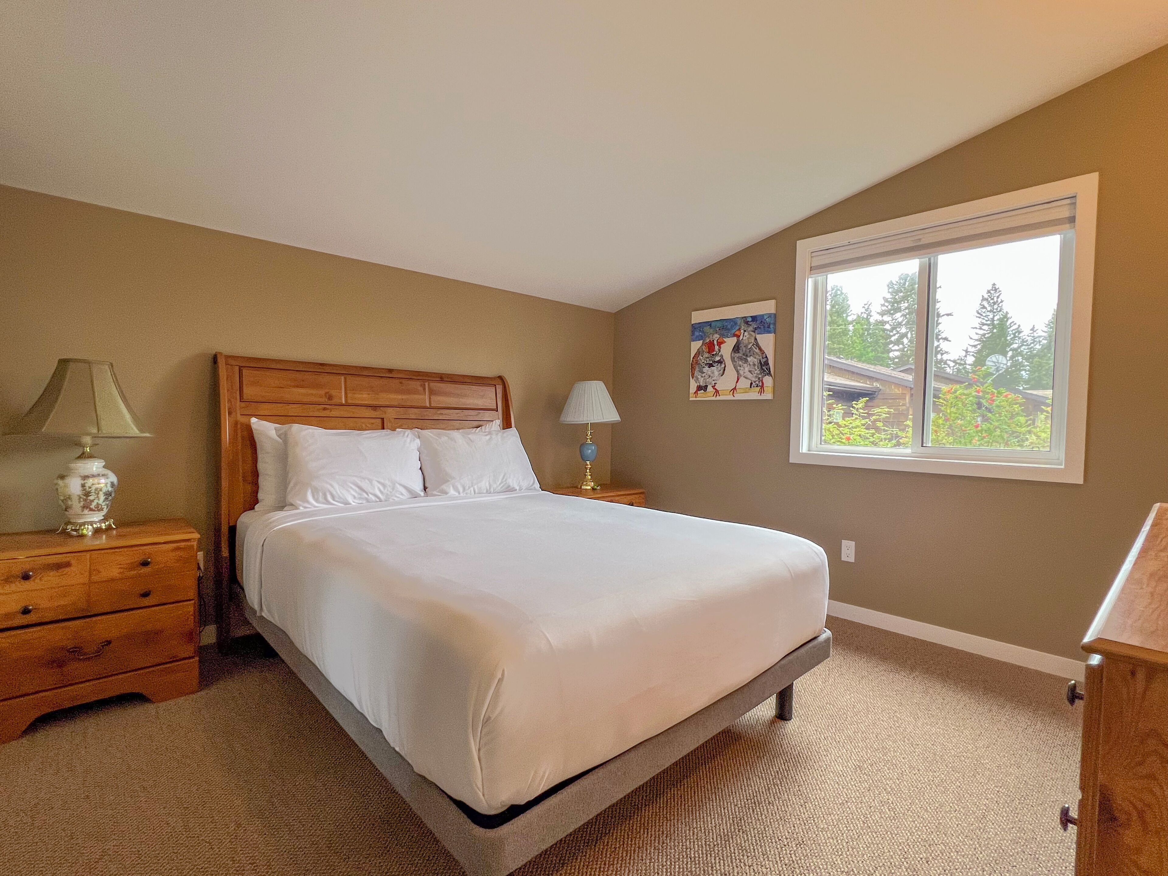 A bedroom with a wooden bed, two bedside tables with lamps, a window with a view of greenery, and a painting on the wall.