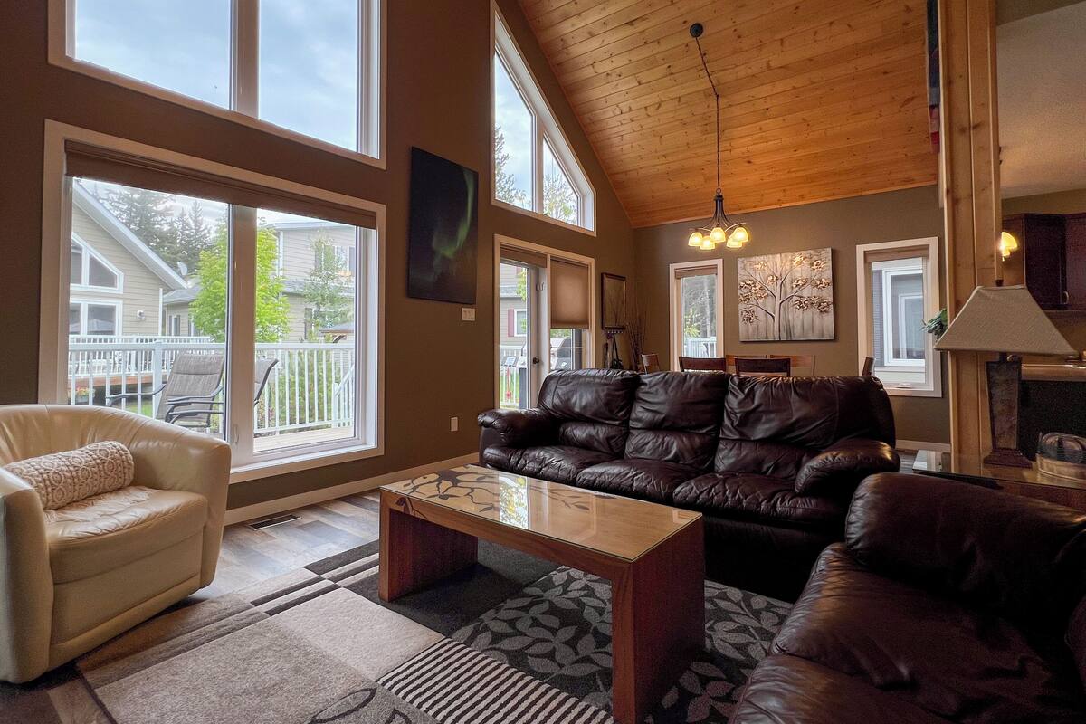 A spacious living room with a high ceiling, large windows, a leather sofa, a coffee table, and a fireplace.