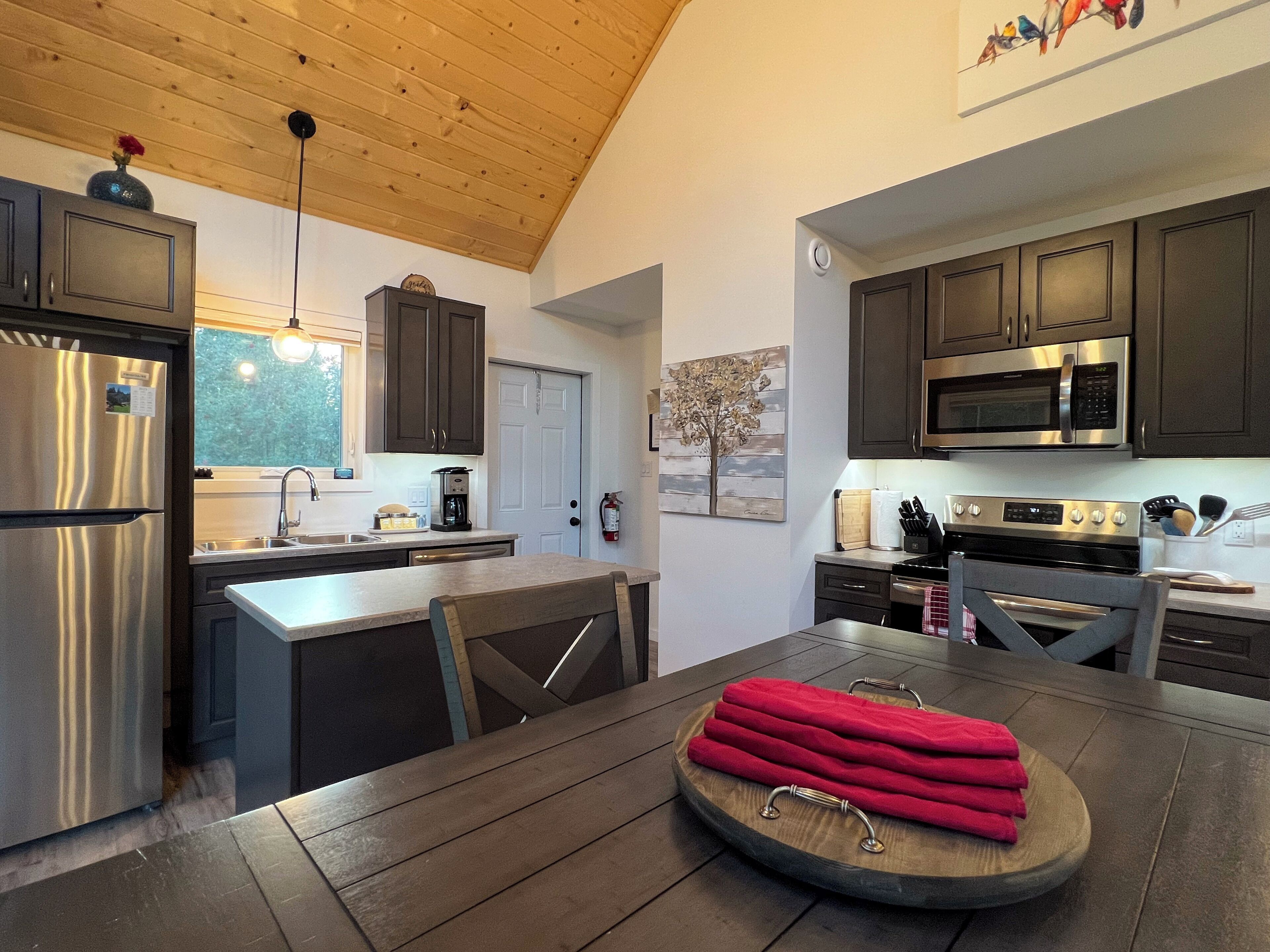 A modern kitchen with dark wooden cabinets, stainless steel appliances, and a wooden dining table with red towels.