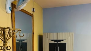 Une pièce avec un plancher en bois, un miroir, une armoire, un bureau avec une chaise et un ventilateur sur pied.