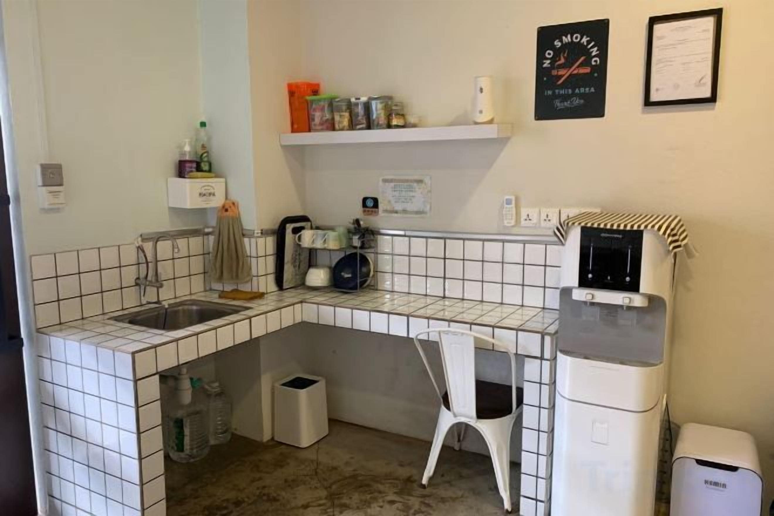A compact kitchen with a sink, a coffee machine, and a microwave. There is a white chair and a trash bin.