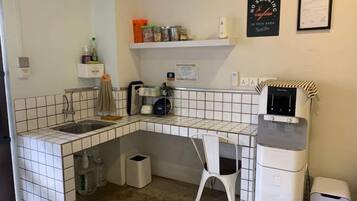 A compact kitchen with a sink, a coffee machine, and a microwave. There is a white chair and a trash bin.
