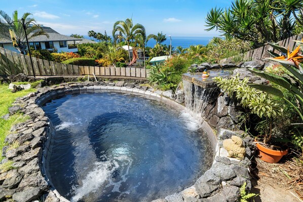 A hot spring surrounded by a wooden fence, tropical plants, and a view of the ocean.