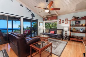 A spacious living room with a leather sofa, a glass coffee table, and a ceiling fan. Large windows offer a view of the ocean.