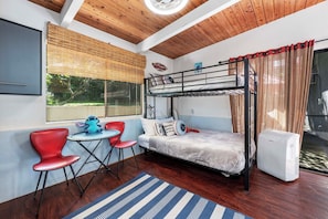 Habitación con literas, una mesita pequeña y dos sillas rojas, una ventana con persianas y un ventilador de techo.