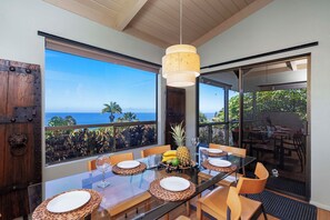 A dining area with a large table set for four, a hanging light fixture, and a view of the ocean through large windows.