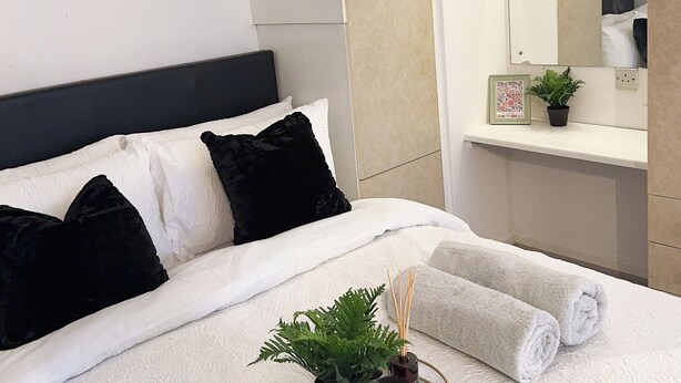 Una cama bien hecha con ropa blanca y almohadas negras, una mesita de noche con una planta y velas, y un espejo grande sobre la cama.