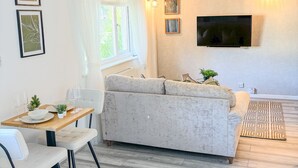 Una sala de estar moderna con un sofá, una mesa de comedor con sillas, un televisor de pantalla plana montado en la pared y una ventana con cortinas.
