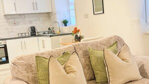 Una sala de estar con un sofá beige, dos almohadas verdes, una mesa de centro y una cocina al fondo.