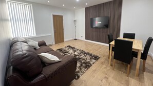 A modern living room with a brown leather sofa, a wooden dining table with black chairs, a flat-screen TV mounted on a striped wall, and a large window with blinds. - Cozy Newcastle Apartment With 2 Bedrooms (Newcastle-upon-Tyne)