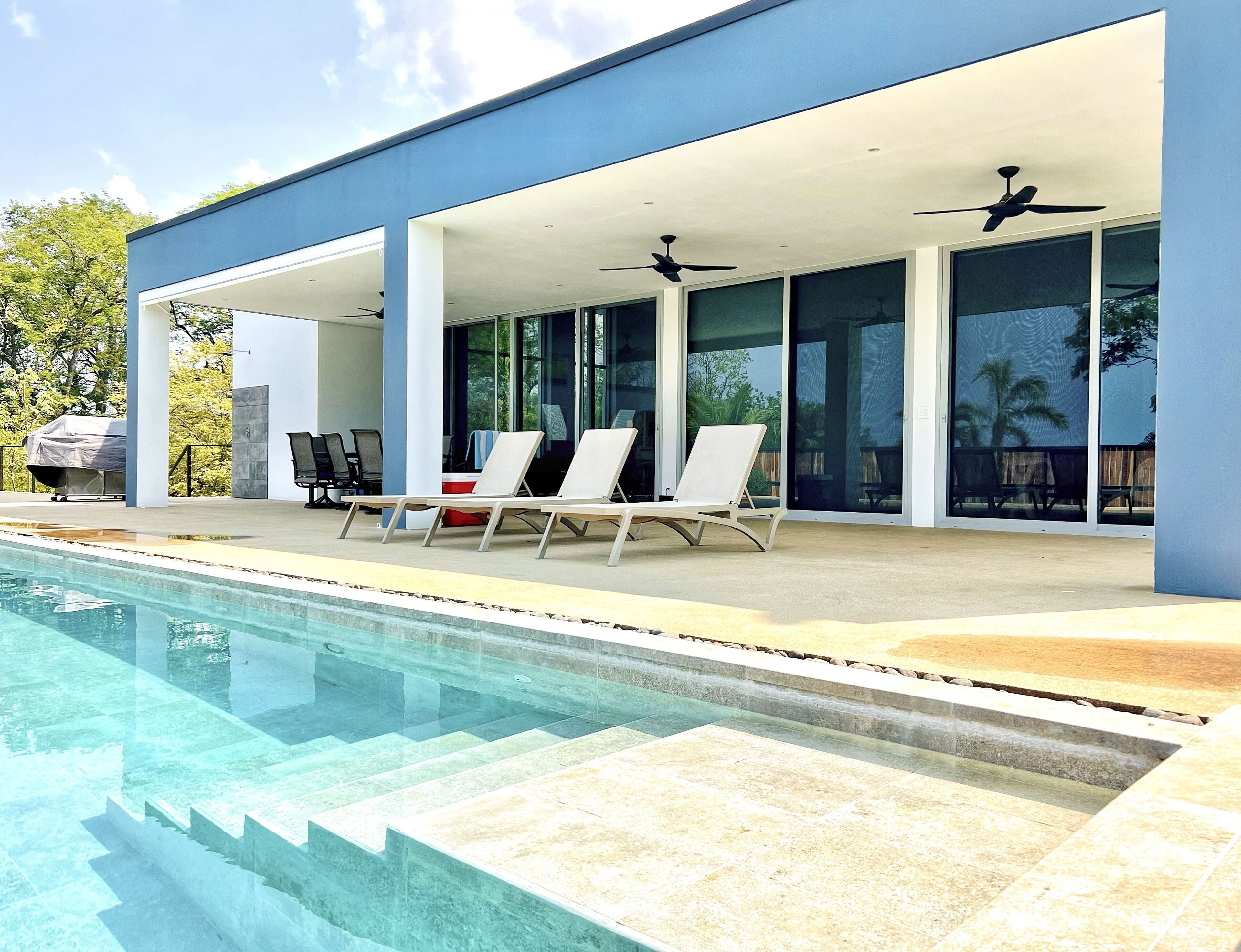 Ein moderner Poolbereich im Freien mit überdachter Lounge, Liegestühlen und freiem Blick auf den Pool.